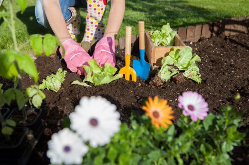 A person's hands, wearing pink gardening gloves, are working in a garden bed with dark, fertile soil, planting young green lettuce plants. In the foreground, a variety of colorful flowers including white, orange, and purple blooms border the garden bed, adding vibrant contrast to the natural tones. Behind the person, there are small wooden garden markers and a blue garden tool partially embedded in the soil, indicating ongoing planting activity. To the left, lush green foliage and additional garden plants are visible, suggesting a well-maintained outdoor space typical of a residential garden in Enfield. The garden area appears neatly organized with defined borders between the flower bed and the surrounding lawn, which is covered in bright green grass. The scene is lit by natural daylight, likely on a clear day, creating a fresh and healthy outdoor environment suitable for gardening services offered by companies like Hedge Trimming Enfield, focusing on planting, lawn care, and garden maintenance.