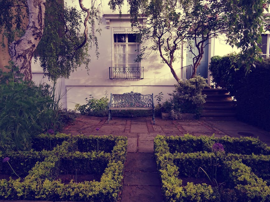A small, carefully maintained front garden featuring a paved patio area with a decorative wrought-iron bench positioned in front of a white house wall. To the left, a large tree with textured bark and