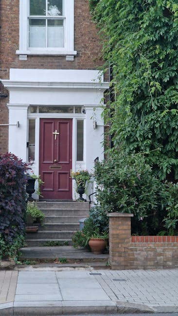 A small, carefully maintained front garden featuring a paved patio area with a decorative wrought-iron bench positioned in front of a white house wall. To the left, a large tree with textured bark and