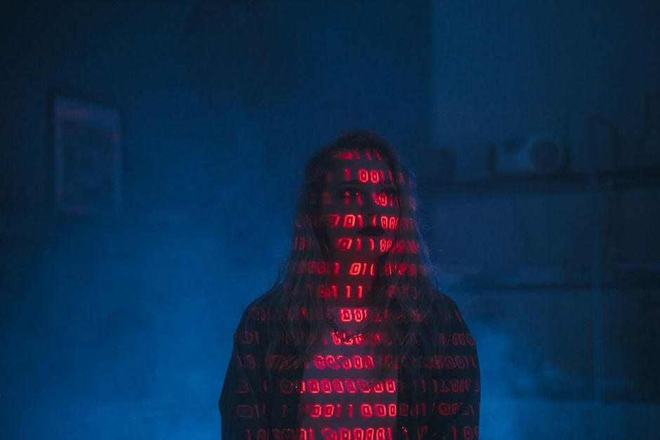 A person standing in a dark room illuminated by red, glowing, floating binary code (ones and zeros) projected onto their face and clothing. The individual has long hair and is dressed in dark attire, 
